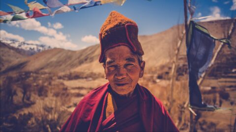 An elderly person with a warm smile, dressed in traditional attire, stands before a scenic mountainous backdrop, flanked by colorful prayer flags fluttering in the breeze.