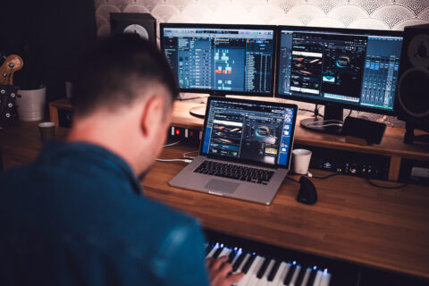 Un sound designer concentré sur la création de pistes sur un ordinateur portable équipé d'un logiciel de station de travail audio numérique, d'un clavier midi et de moniteurs de studio dans une configuration de studio à domicile.
