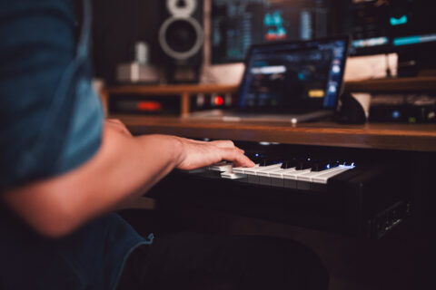Sound designer at an electronic keyboard with a focus on the hands playing keys, set against a background of a home studio with recording equipment.