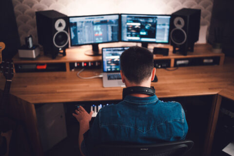 Un sound designer de dos, travaillant dans un studio d'enregistrement avec plusieurs écrans d'ordinateur et des haut-parleurs sur un bureau en bois. Il porte une chemise en jean et a un casque autour du cou.