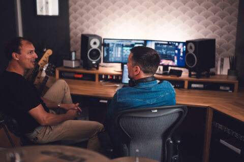 Two sound designers in a modern home studio setup engaged in a music production session with professional audio equipment in the background.