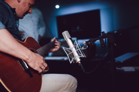 A musician strums an acoustic guitar with focus, as the microphone awaits to capture the melody in a studio setting.