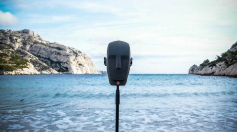 Un microphone tête binaural placé sur un pied, devant une plage avec des montagnes en arrière-plan. Le ciel est partiellement nuageux.
