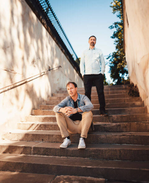 two men on a rock stairs outside. one is seated the other is standing on his feet.