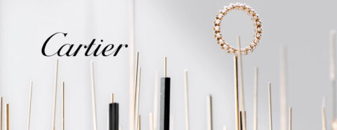 Un anneau en or avec des pierres précieuses, monté sur un support, avec le logo "Cartier" en noir sur un fond clair parsemé de tiges dorées et noires.