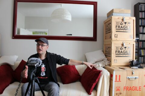 Alexandre dubosc assis sur un canapé, avec des boîtes en bois marquées "Fragile" en arrière-plan.