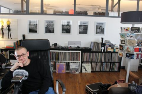 Laurent Brett assis dans un studio de musique avec une collection de vinyles en arrière-plan.