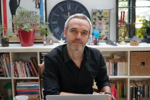 Olivier Bal assis devant une étagère avec des livres et des décorations, regardant vers la caméra.