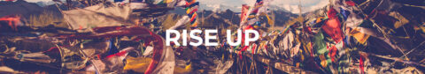 L'image montre une bannière avec des drapeaux de prière colorés flottant dans le vent, typiques de la région de l'Himalaya. En arrière-plan, on aperçoit des montagnes majestueuses sous un ciel clair. Au centre de l'image, le texte "RISE UP" est écrit en lettres blanches majuscules, attirant immédiatement l'attention.