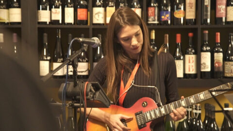 Musicienne jouant de la guitare électrique devant des étagères remplies de bouteilles de vin.