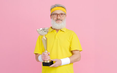 L'image montre un homme âgé portant un bandeau et un polo jaune, tenant un trophée en or. Il a une barbe blanche et porte des lunettes. Le fond est rose vif, créant un contraste frappant avec la tenue jaune de l'homme.