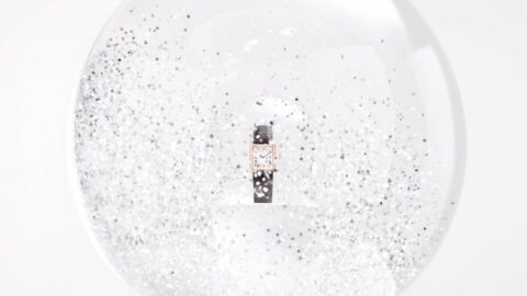 Une montre avec un bracelet en cuir noir et un cadran rectangulaire, présentée dans une sphère transparente remplie de paillettes argentées.